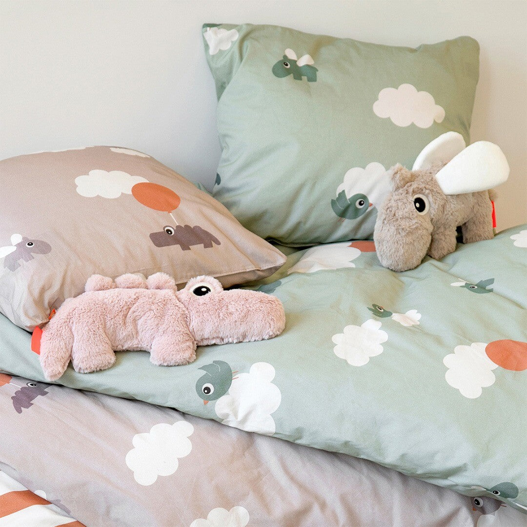 Twee Done by Deer pluchen knuffelnijlpaarden, een zandkleurige en een roze, liggen op een bed met beddengoed met wolkenpatroon.