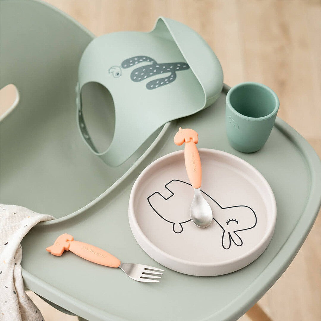 Een Done by Deer babyvoedingsset met een beige bord, oranje bestek, een groene slab en beker op een kinderstoel.