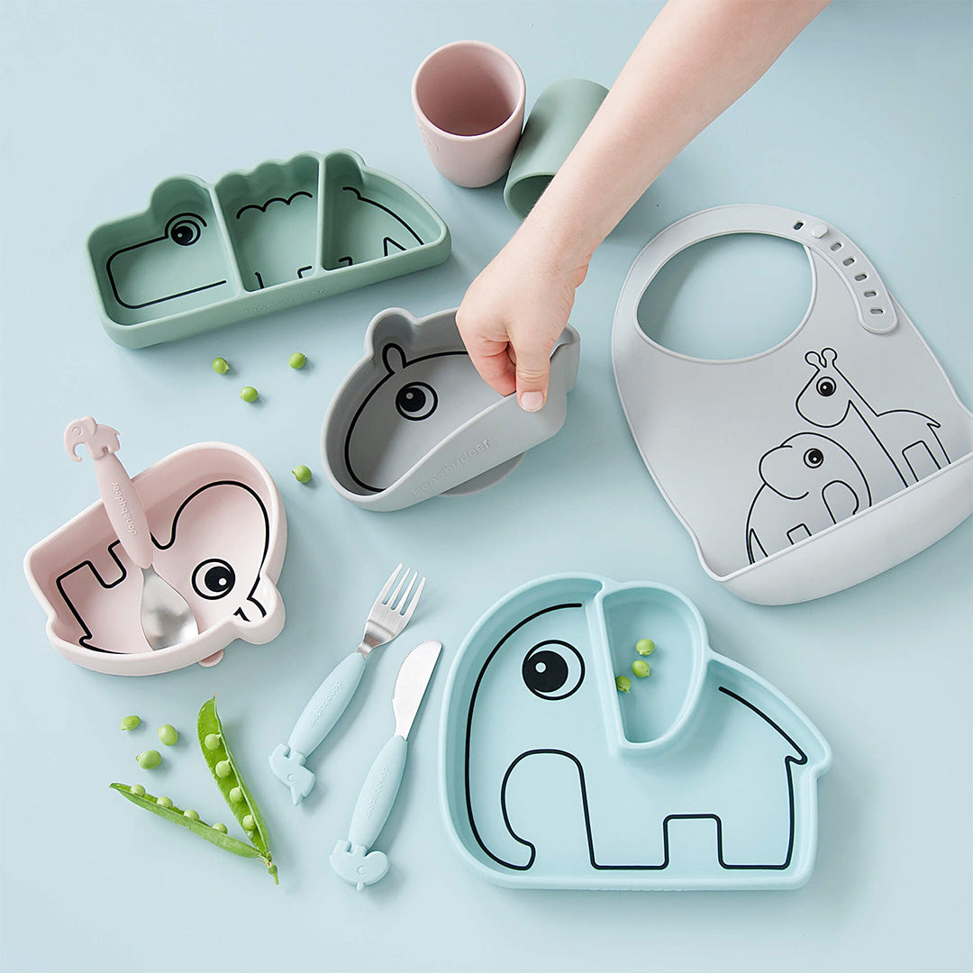 Een collectie done by deer baby voedingsbenodigdheden met onder andere een groen krokodil snackbord, een roze olifantenbord en een grijze slab.