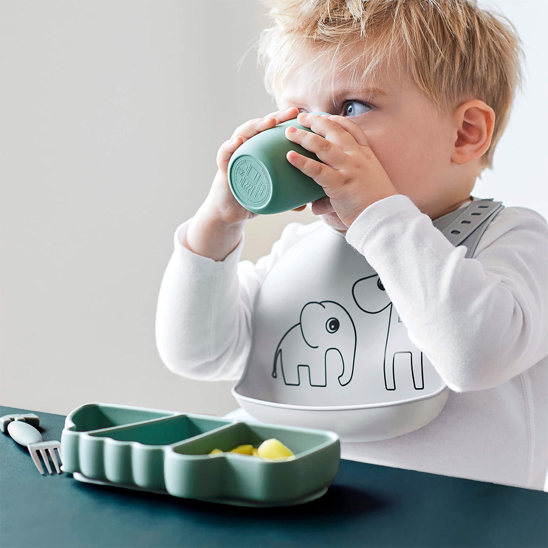 Een peuter drinkt uit een groene beker, met op de voorgrond een groen done by deer krokodil snackbord met fruit.