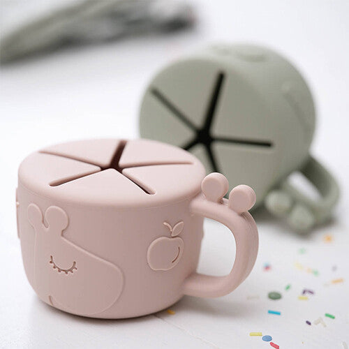 Done by Deer roze siliconen snackdoosje met giraffe en fruit, met een groen exemplaar op de achtergrond.