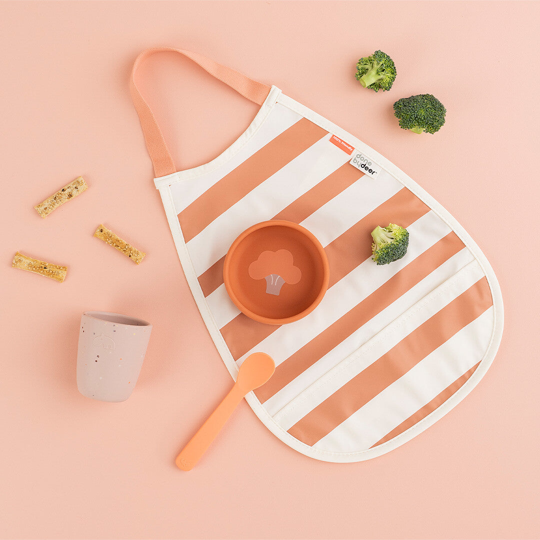 Done by Deer gestreept baby slabbetje in papaya kleur, liggend naast een kommetje, lepel, beker en broccoli op een roze ondergrond.