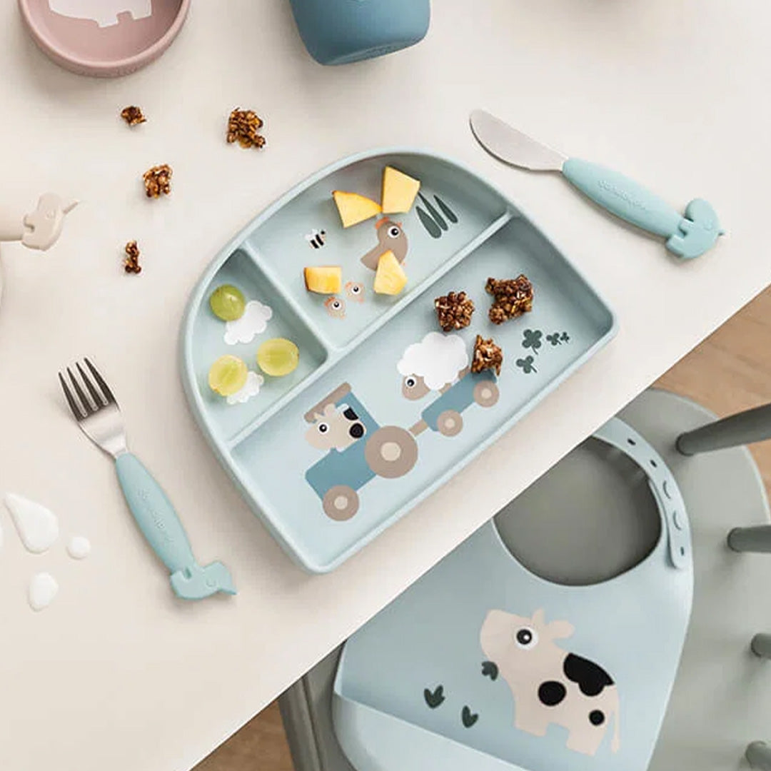 Done by deer siliconen babybord met drie vakjes, gevuld met fruit en granola, op een tafel met een bijpassend slabbetje.