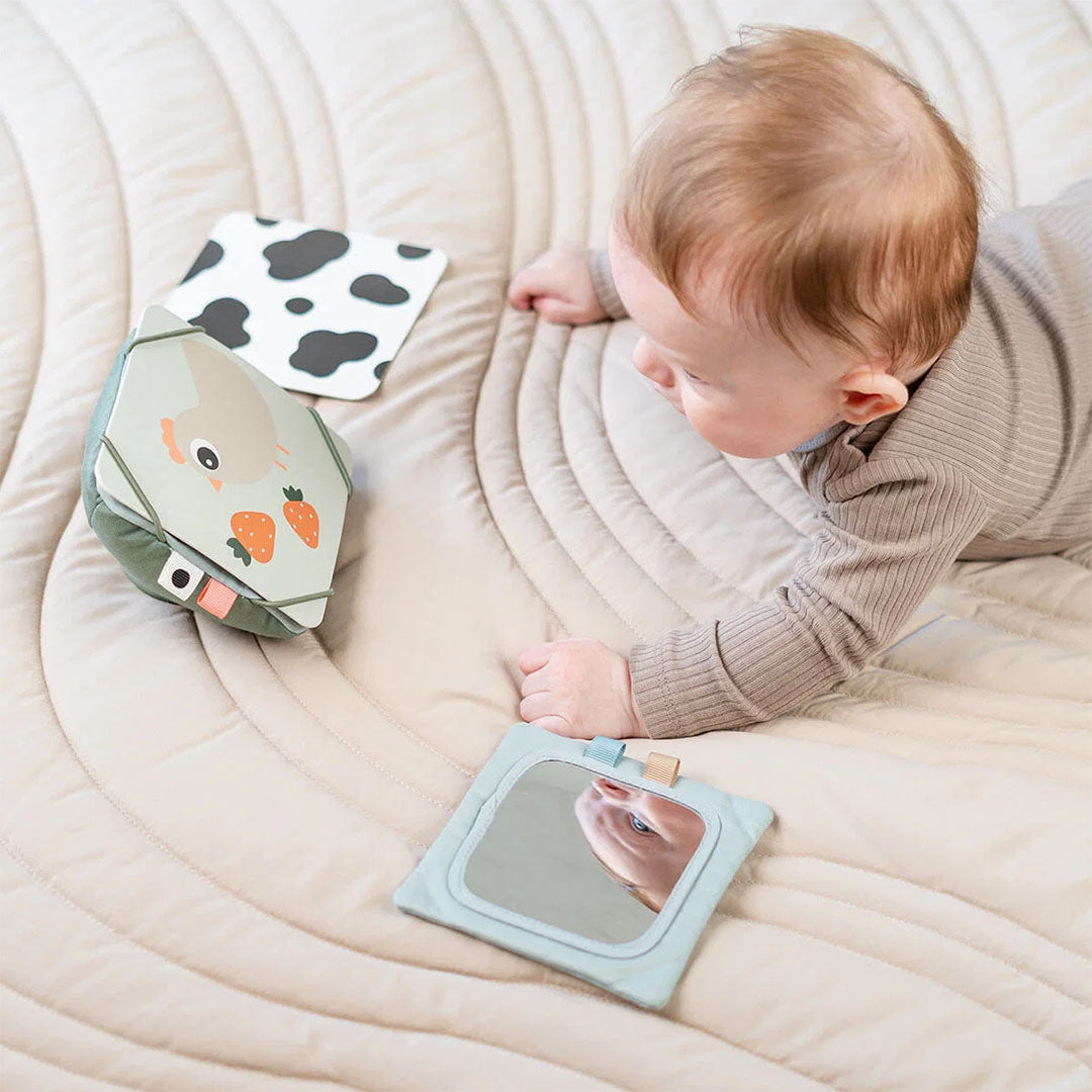 Een baby ligt op een mat en kijkt naar de done by deer activiteitenkaartenset met spiegel en koeienprint.