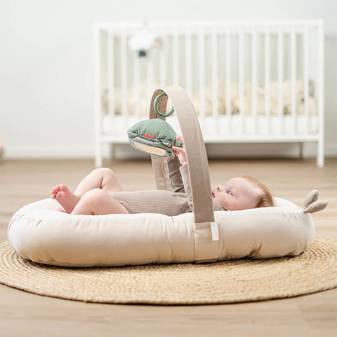 Een baby ligt in een done by deer babygym en kijkt naar een hangend groen speeltje.