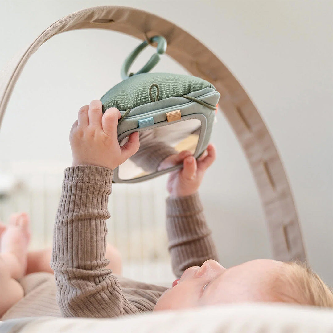 Babyhandjes houden een done by deer activiteitenkaartenset met spiegel vast, hangend aan een babygym.