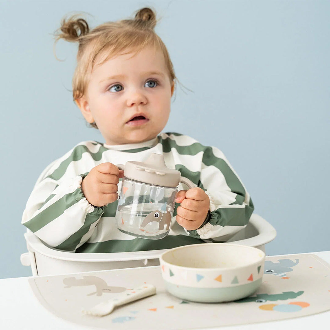 Een baby in een kinderstoel met een gestreepte slab, die een drinkbeker vasthoudt naast een kinderkom.