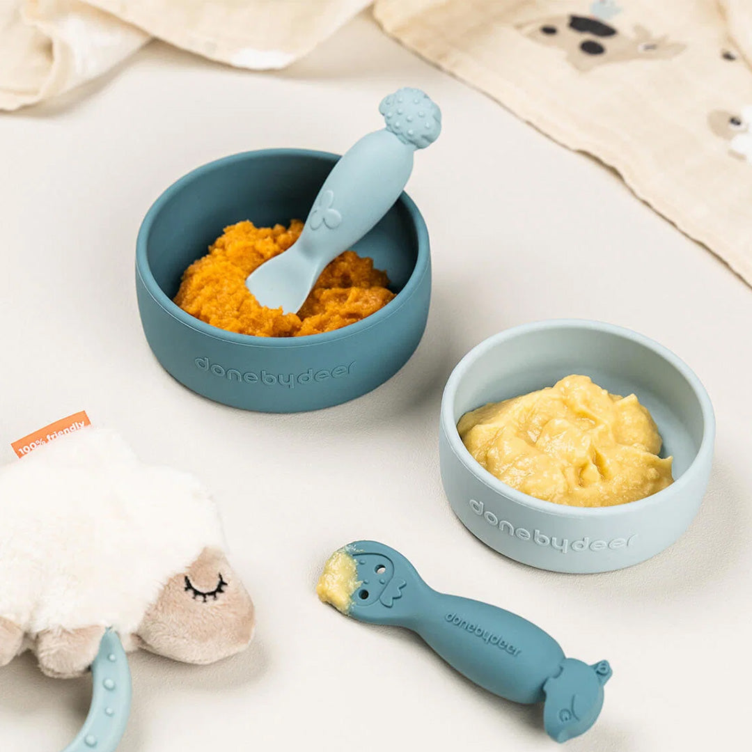 Twee Done by Deer siliconen kinderkommen met babyvoeding en bijpassende lepels, naast een schapenrammelaar.