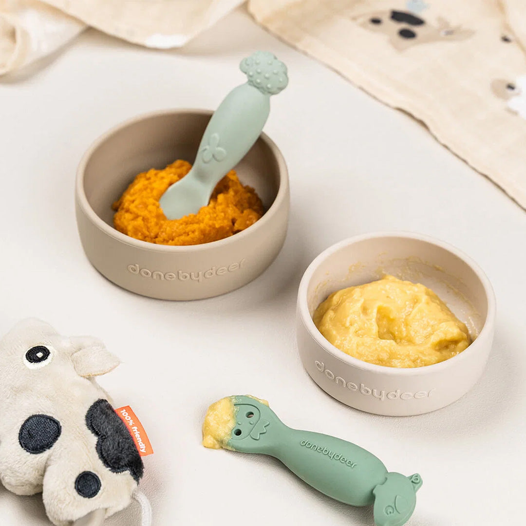 Twee Done by Deer kinderkommen met gepureerd eten en twee groene siliconen lepels.