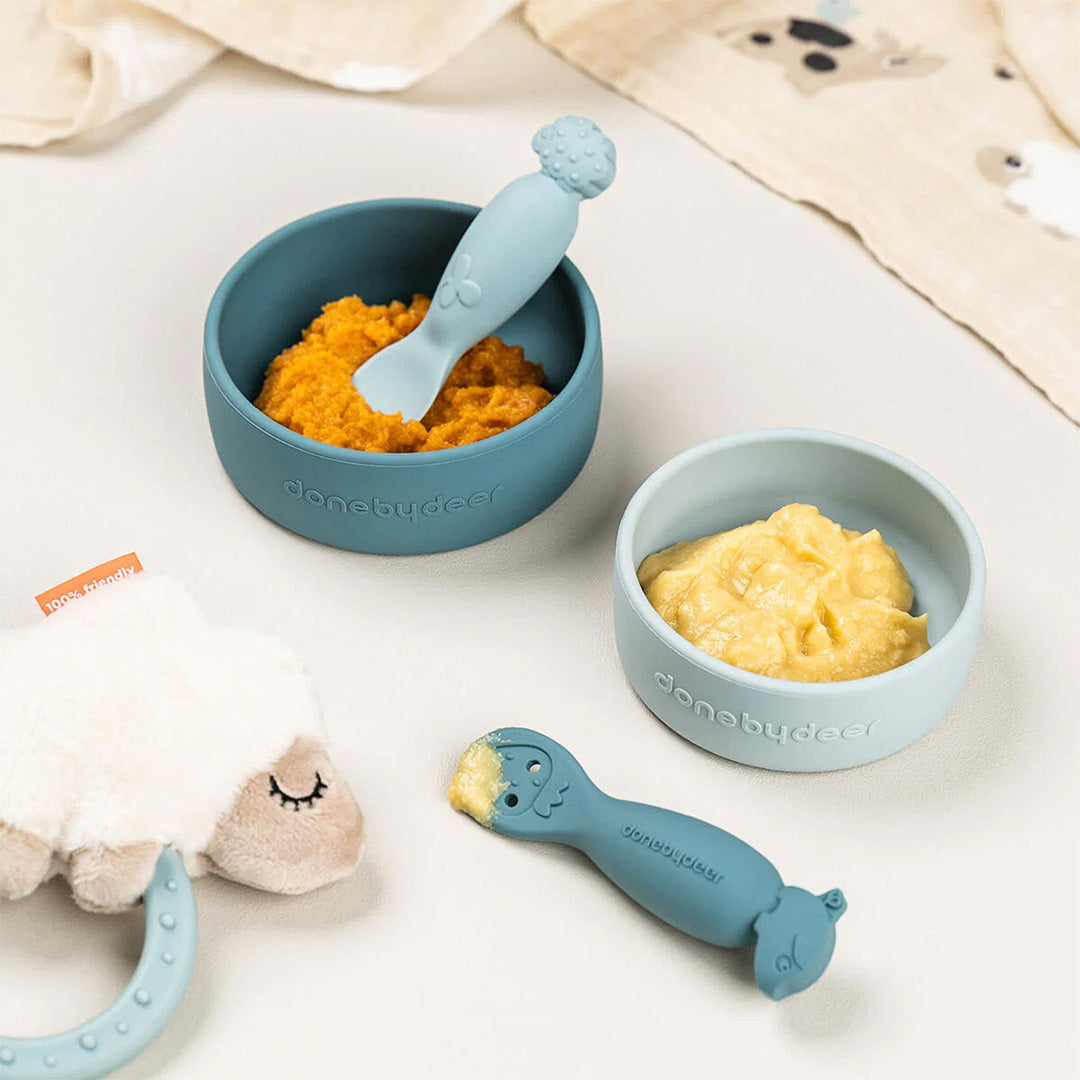 Twee done by deer blauwe kinderlepels met dierenhoofden in en naast kommen met babyvoeding.