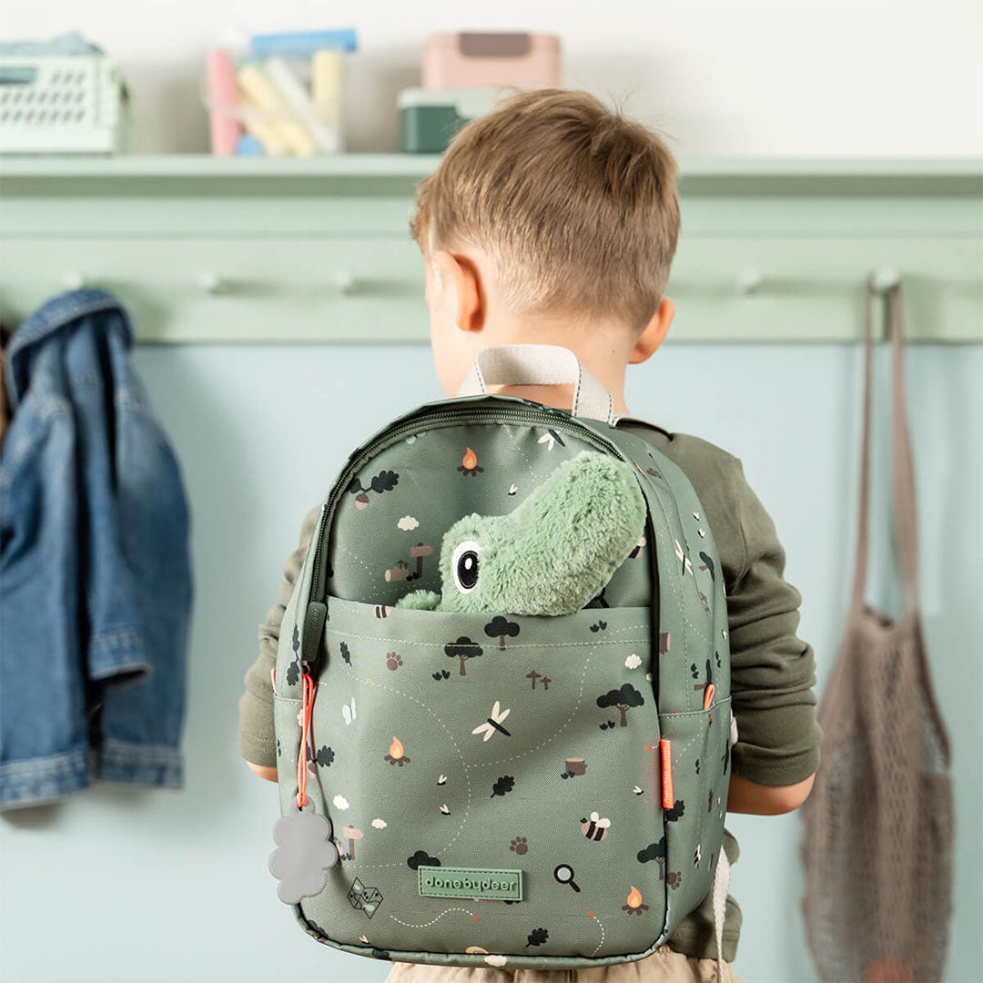 Een kind draagt de Done by Deer groene kinderrugzak met 'tiny trails' patroon, waaruit een knuffelkrokodil steekt.