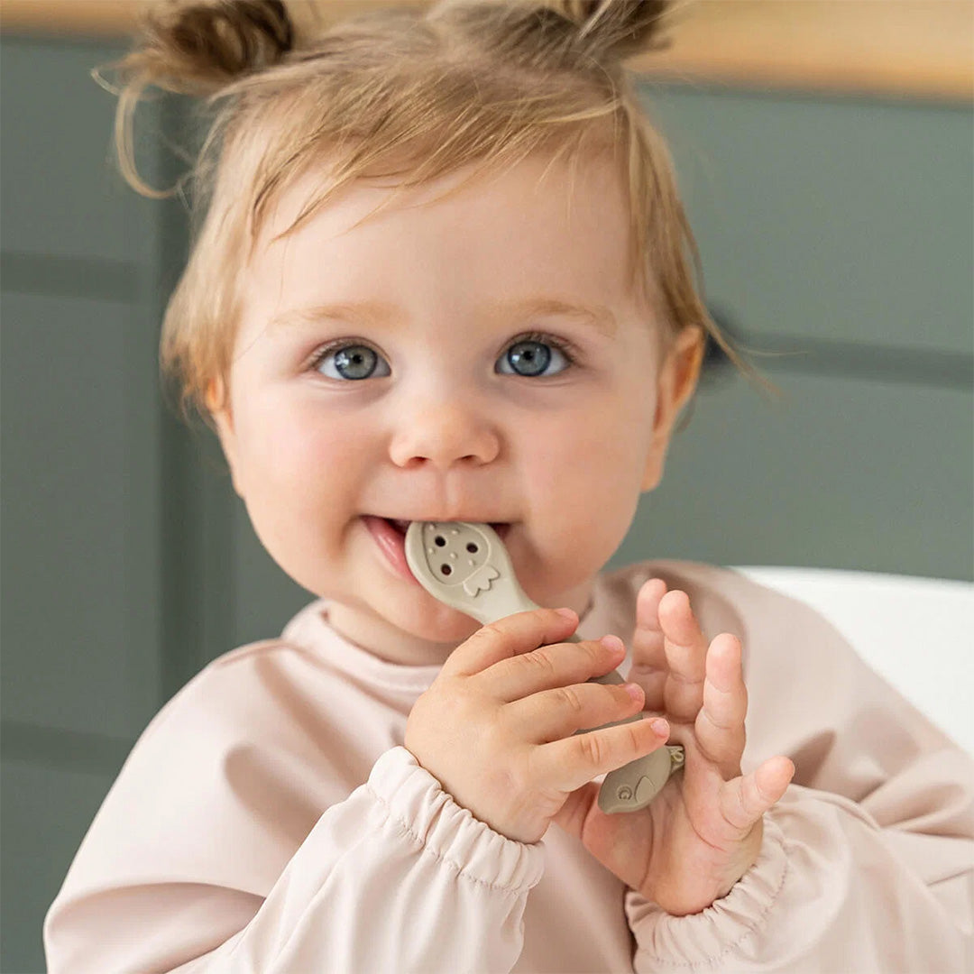 Een lachende baby met een roze slabber houdt een beige kinderservies lepel in de mond.