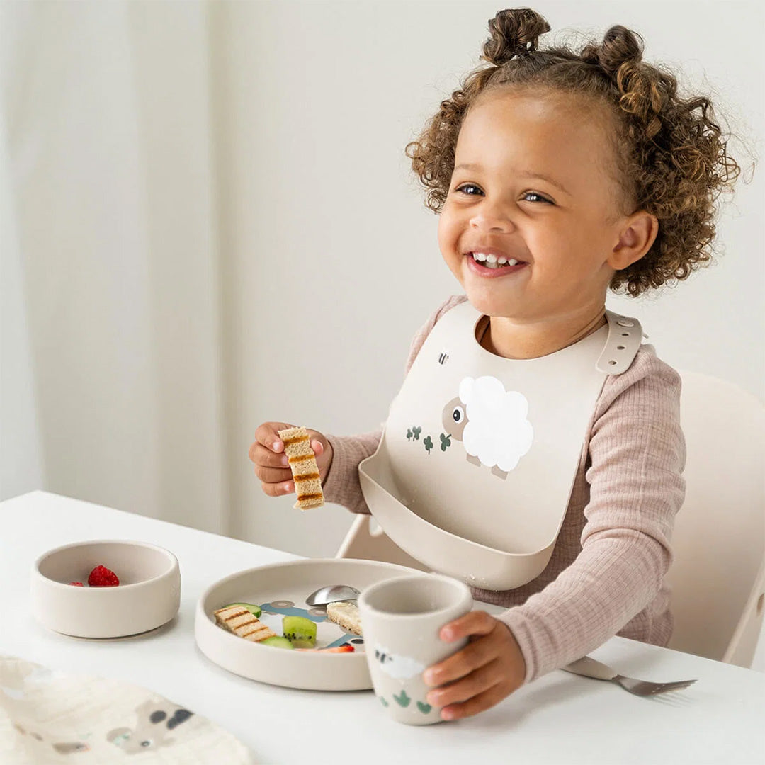 Lachende peuter met krullen draagt een done by deer siliconen slab en eet van het kinderservies.