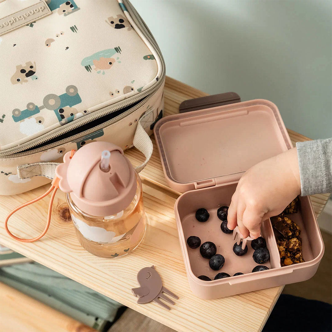 Een kinderhand pakt bosbessen uit een open done by deer roze kinderlunchbox, naast een waterfles en lunchtas.