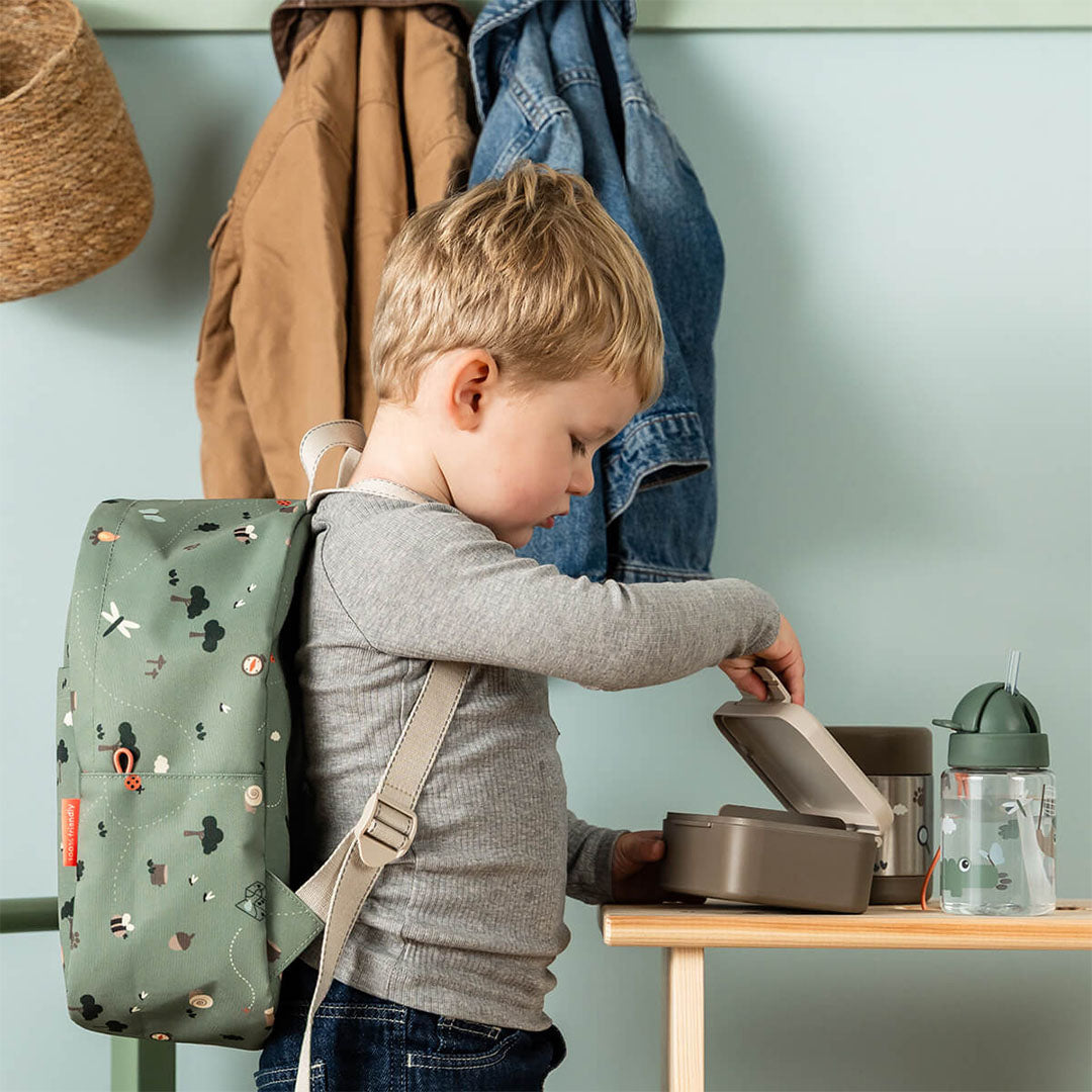 Een kind met een rugzak opent de Done by Deer kinder lunchbox op een tafel naast een drinkfles.
