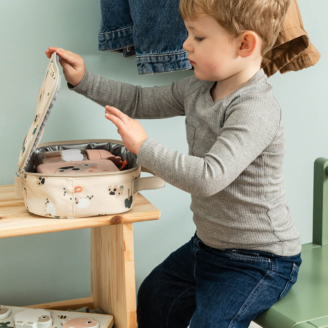 Een jongen opent de beige Done by Deer lunchtas met Tiny Farm patroon, waarin een roze lunchbox zichtbaar is.