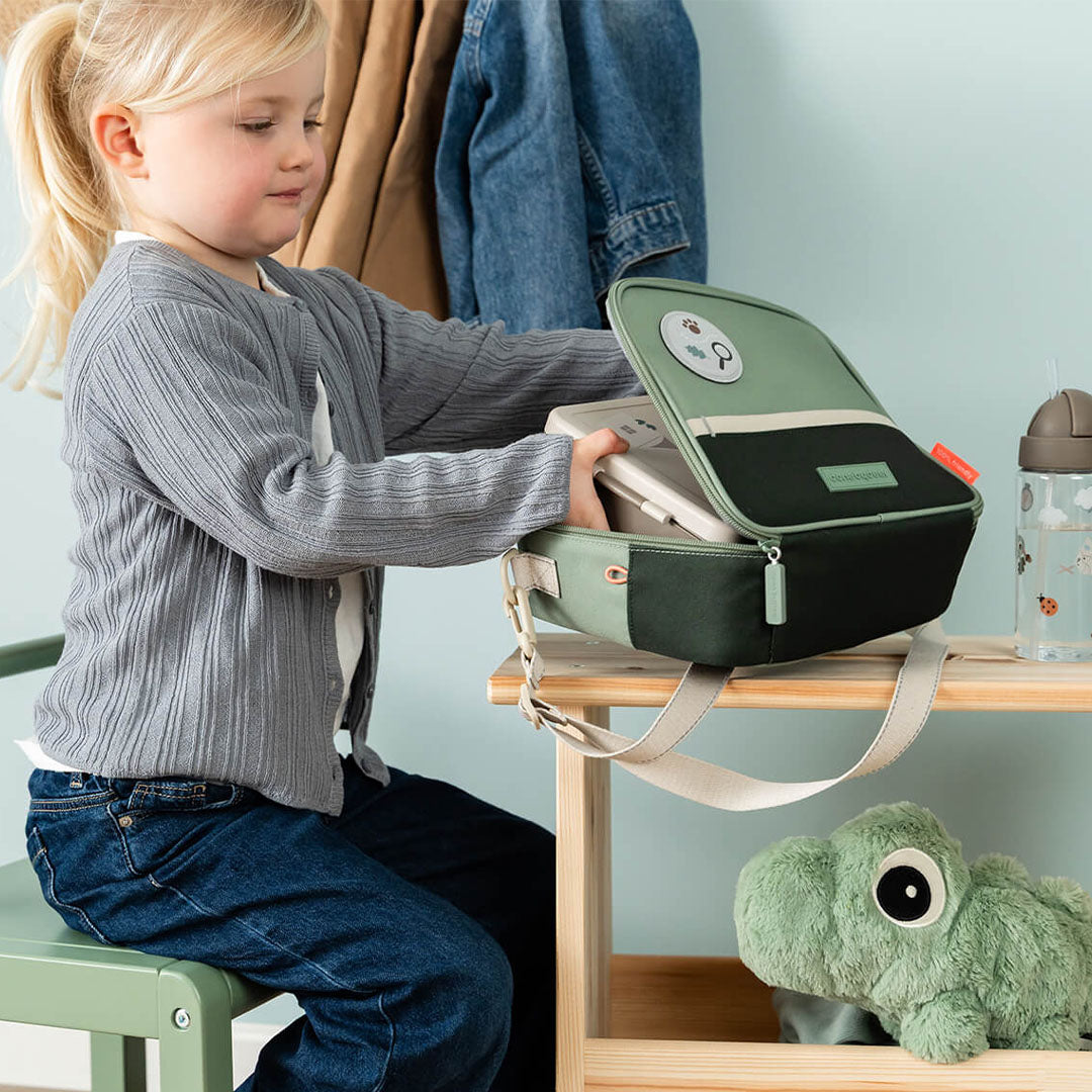 Een kind opent de done by deer groene lunchtas met dierenthema op een houten krukje.