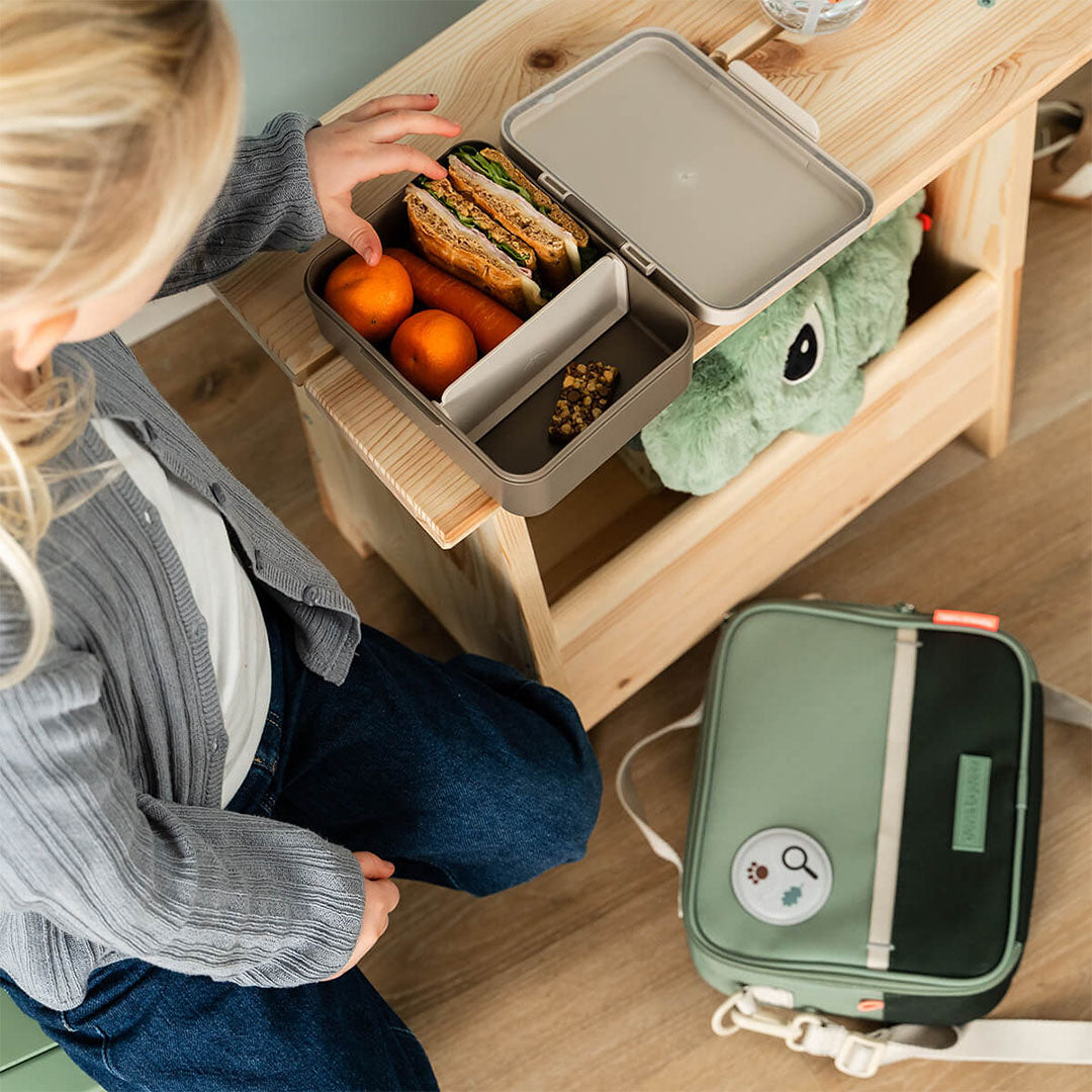 Een kind pakt een lunchbox met broodjes en fruit uit de open done by deer groene lunchtas.