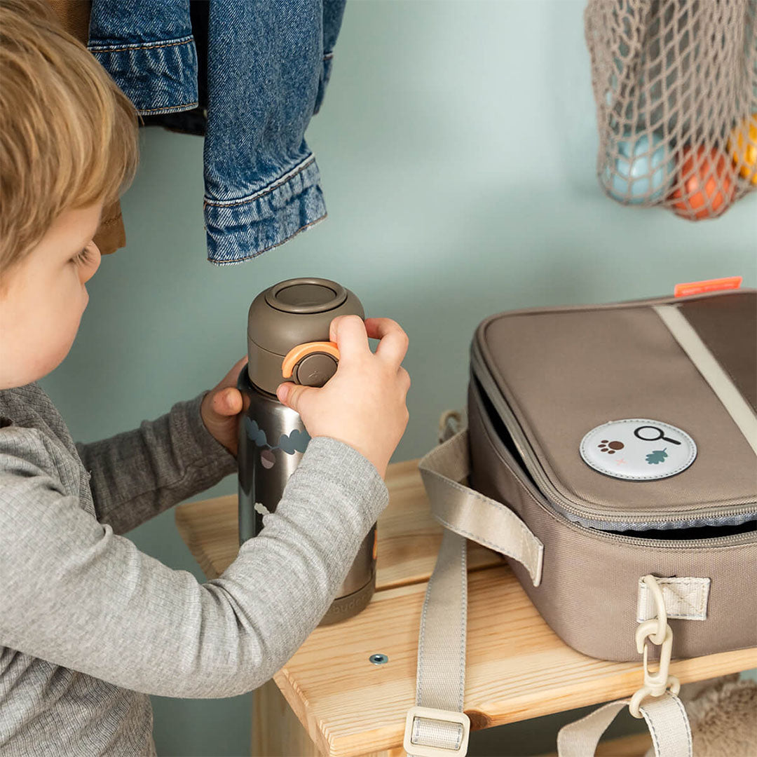 Een kind houdt een thermosfles vast naast de Done by Deer Tiny Trails lunchtas op een houten bankje.