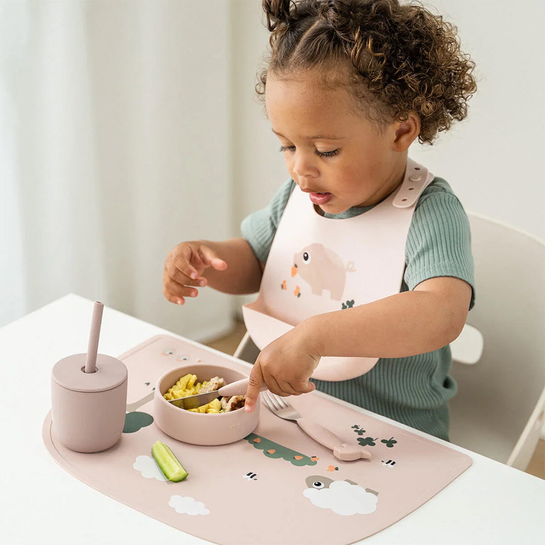 Een kind eet aan tafel met een done by deer roze siliconen placemat en bijpassende voedingsset met Tiny Farm thema.