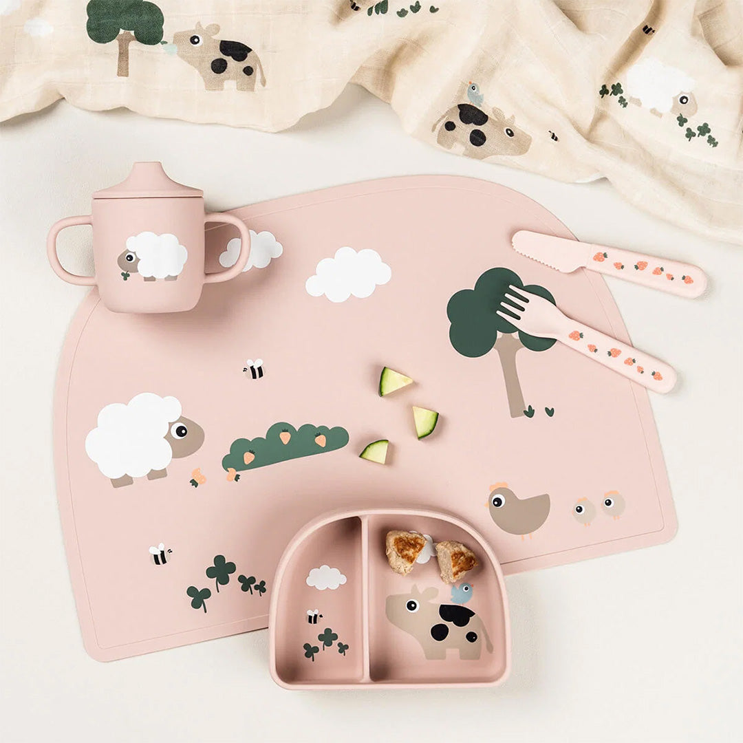 Een done by deer roze siliconen placemat met Tiny Farm dierenpatroon, samen met een bijpassende beker, bestek en verdeeld bord.