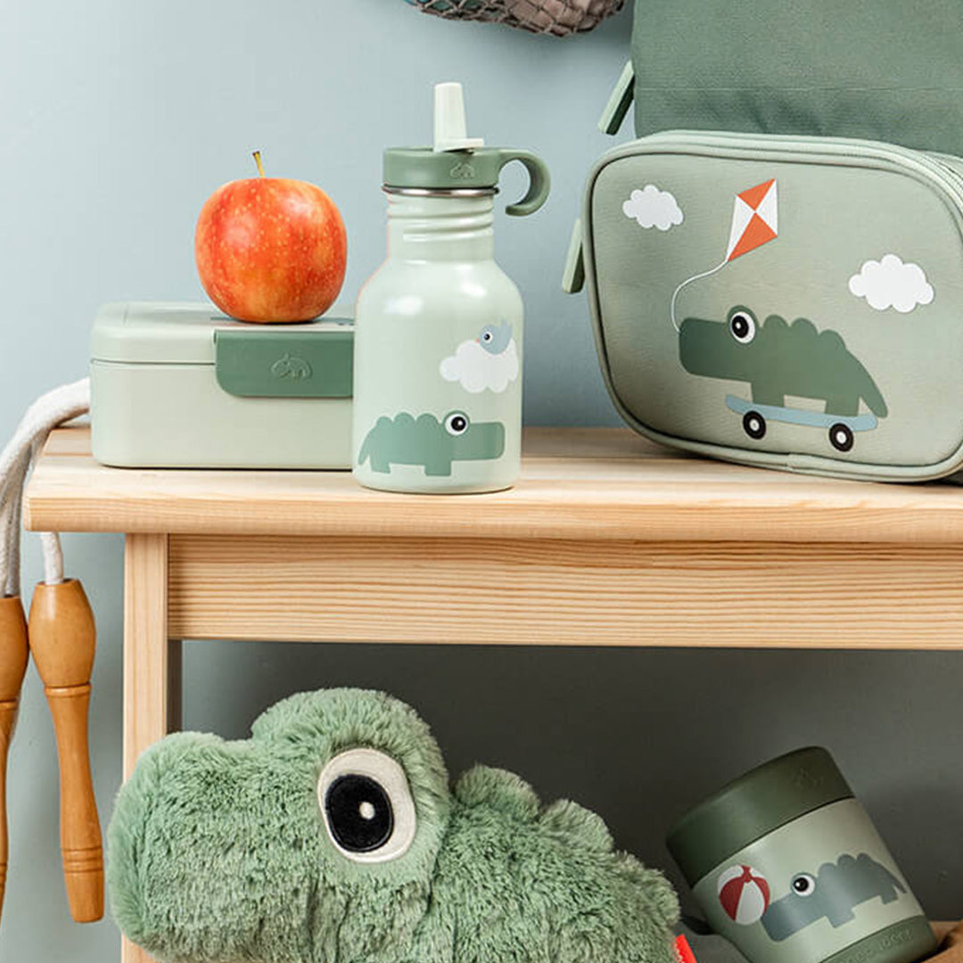 Done by Deer groene drinkfles, lunchbox, lunchtas en knuffelkrokodil op een houten plank.