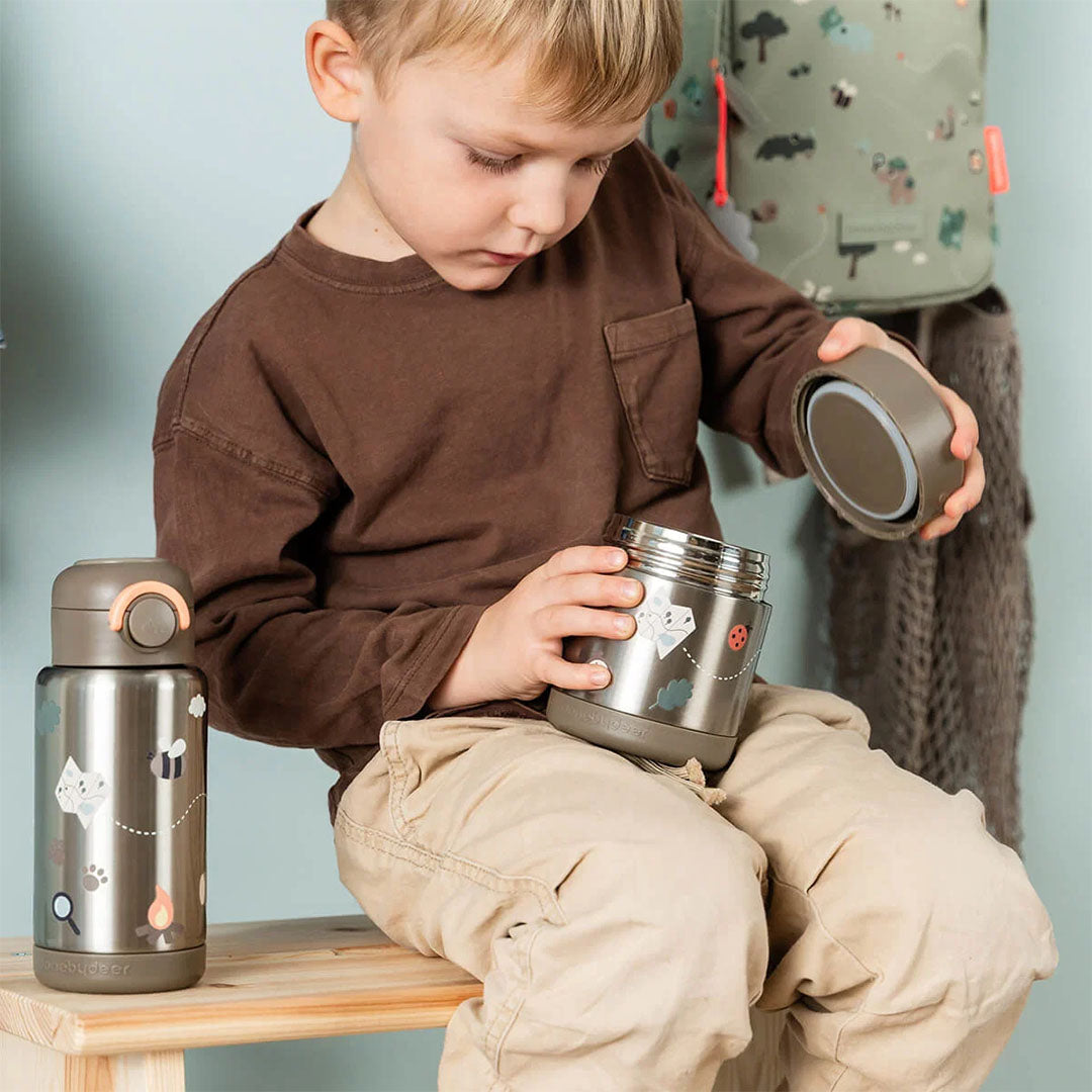 Een kind opent de Done by Deer roestvrijstalen voedselpot met Tiny Trails thema, met een bijpassende thermosfles ernaast.