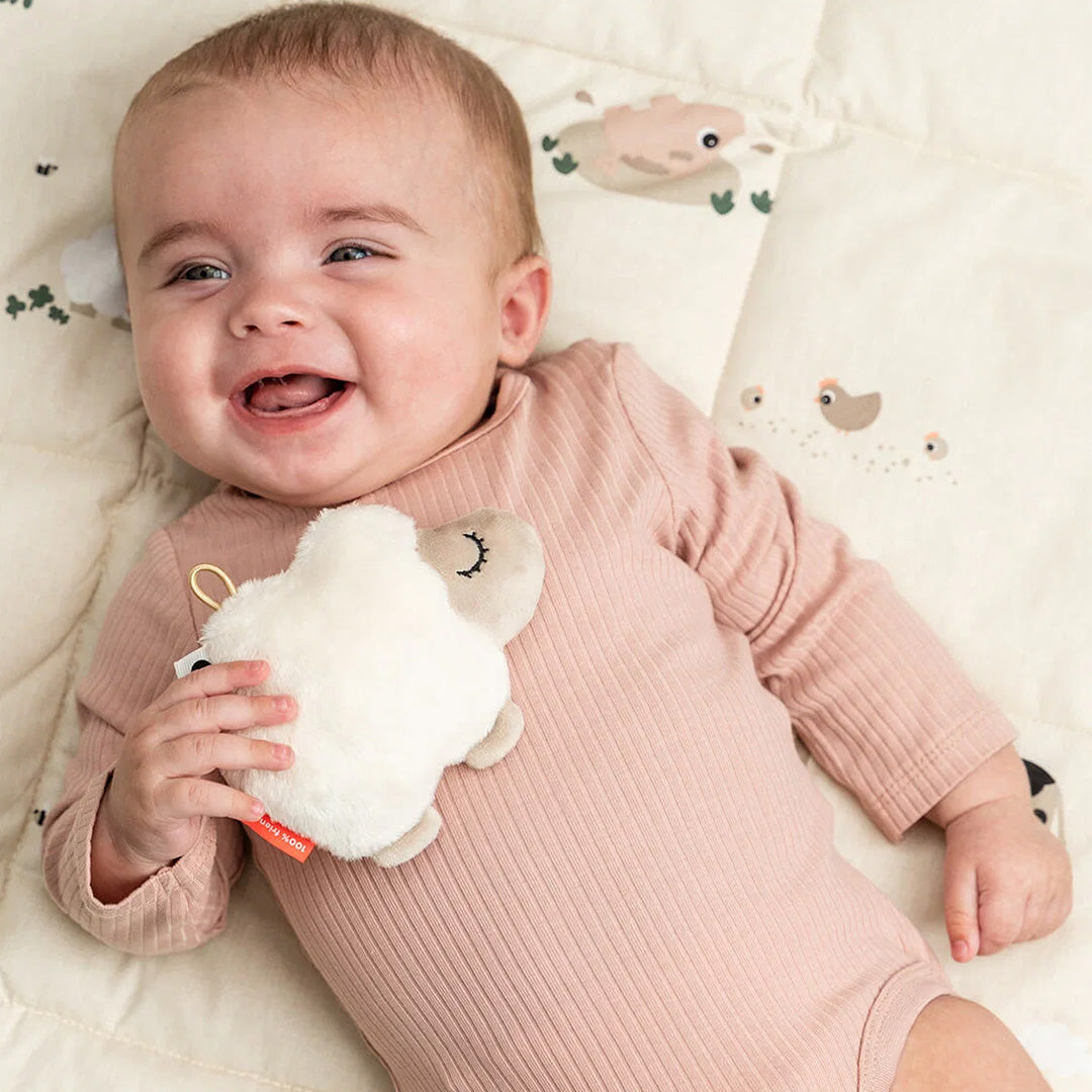 Een lachende baby houdt de Done by Deer schaap rammelaar vast op een speelkleed.