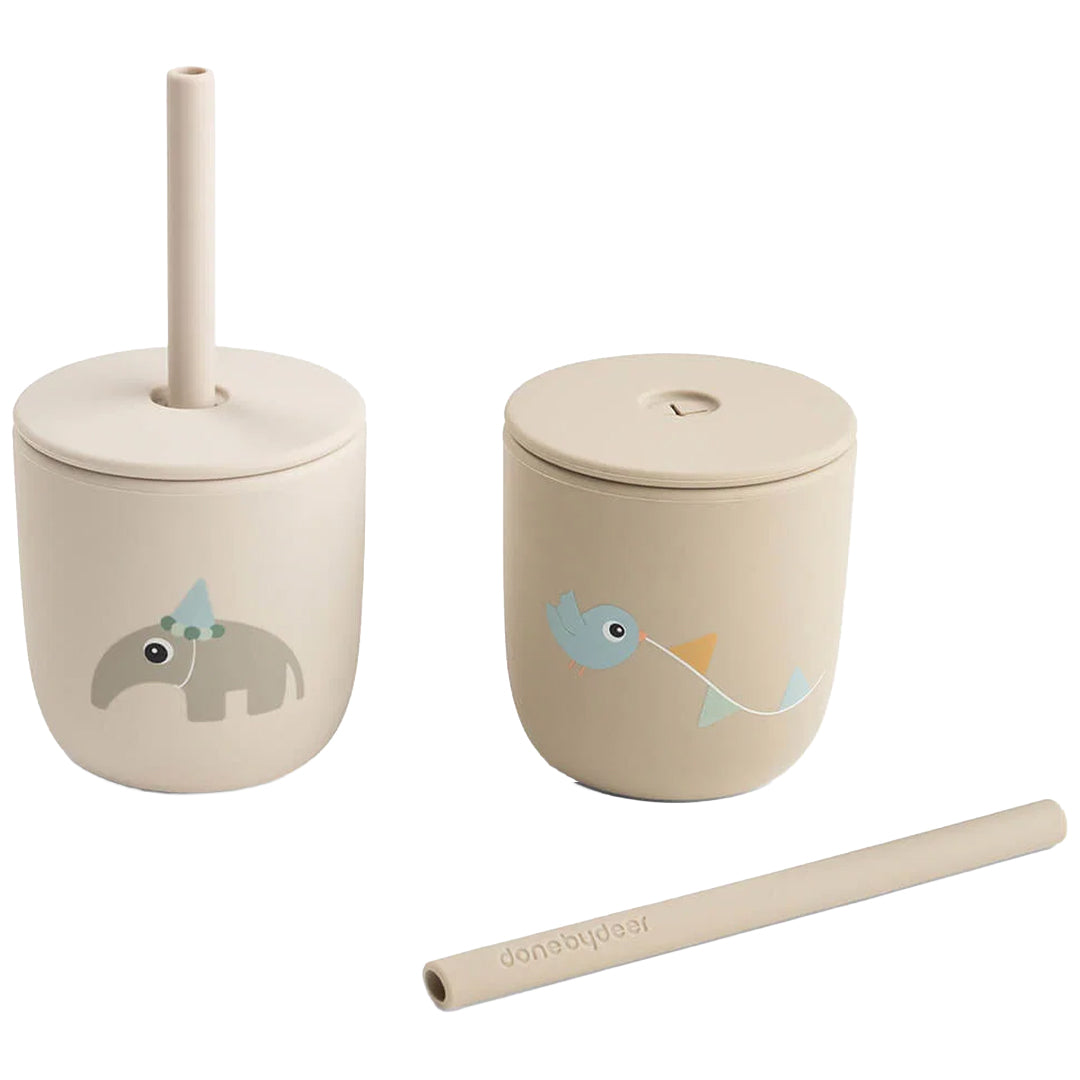 Twee Done by Deer beige kinderbekers met rietjes, waarvan één rietje los ernaast ligt.