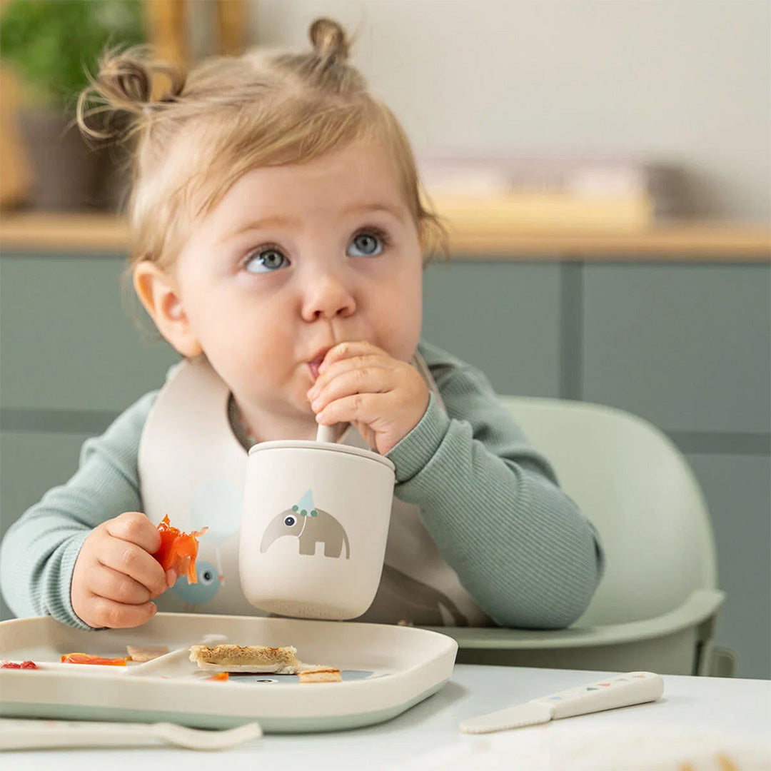 Een baby drinkt uit een Done by Deer beige kinderbeker met olifant en rietje aan een eettafel.