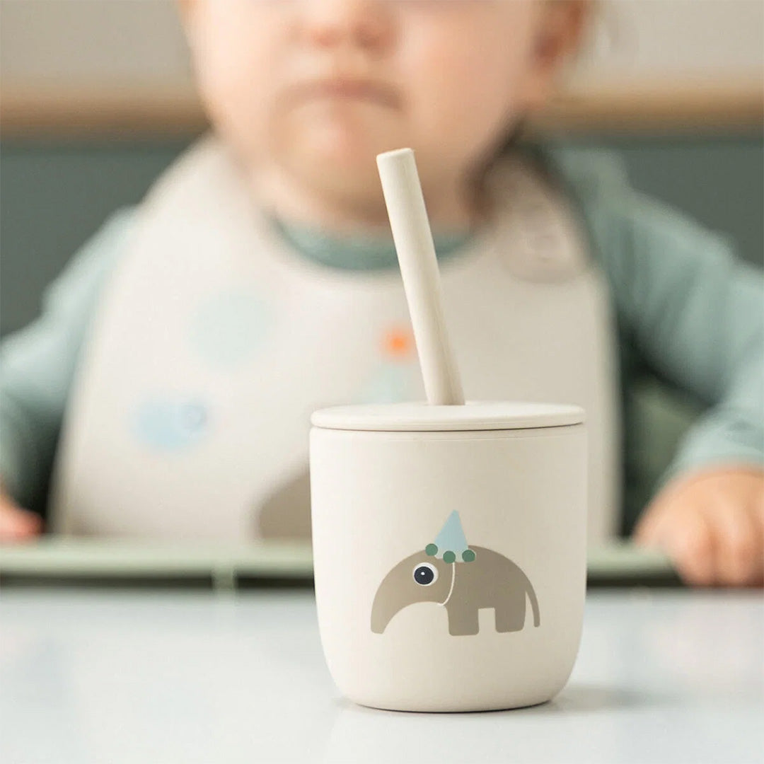 Close-up van een Done by Deer beige kinderbeker met olifant en rietje, met een baby op de achtergrond.