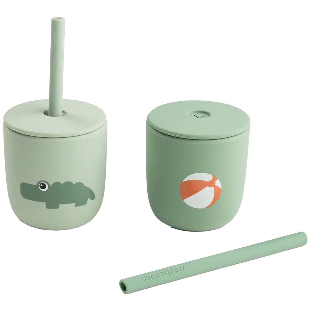 Twee Done by Deer groene kinderbekers met rietje, één met een krokodil en één met een strandbal, met een los rietje ervoor.