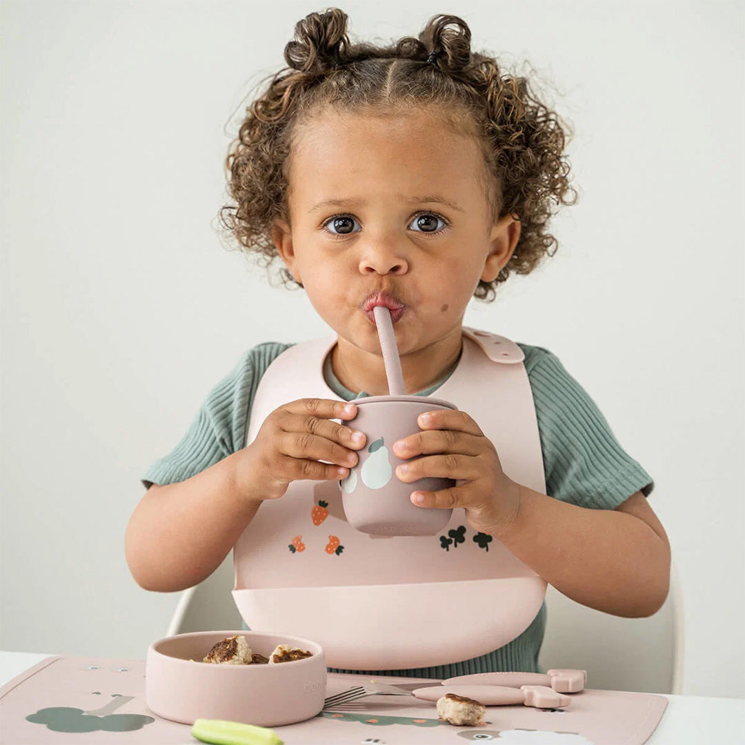 Een kind drinkt uit een roze done by deer kinderbeker met rietje en perenpatroon.