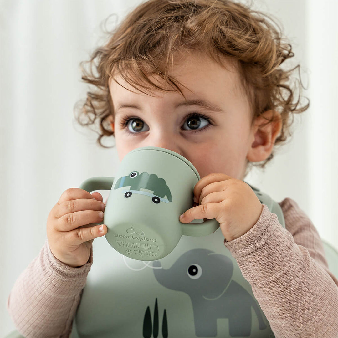 Een baby drinkt uit de done by deer groene tuitbeker met krokodil op skateboard.