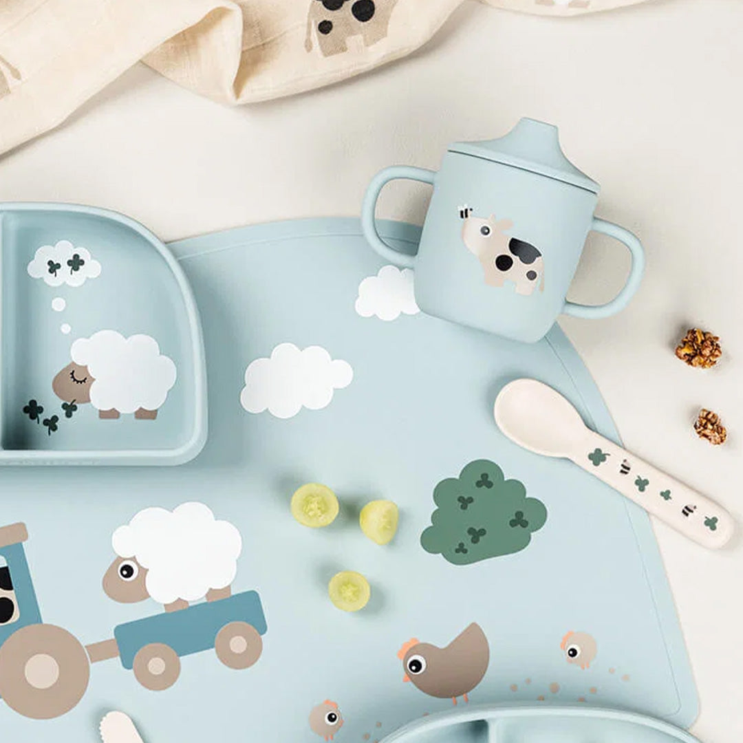 Done by deer siliconen babytuitbeker op een placemat met een trein en schapen, met een lepel en wat eten.