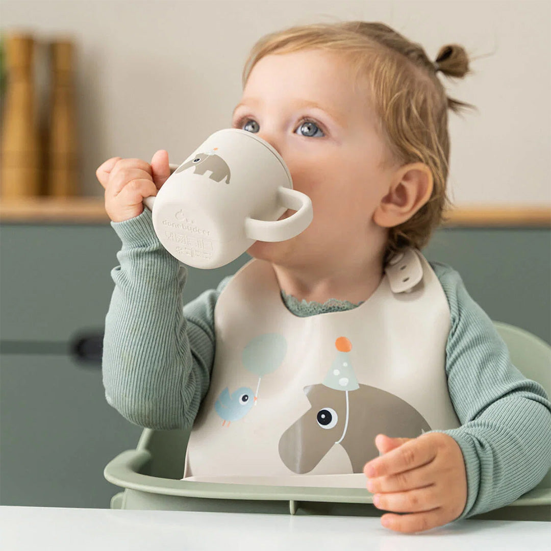 Een baby draagt het done by deer siliconen slabbetje met olifant en vogel terwijl het drinkt uit een beker.