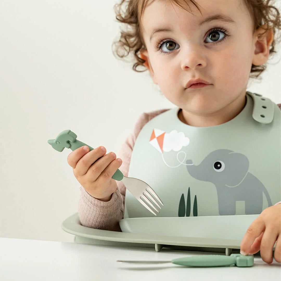 Een baby draagt een Done by Deer groen siliconen slabbetje met olifant en houdt een groen kinderbestek vast.
