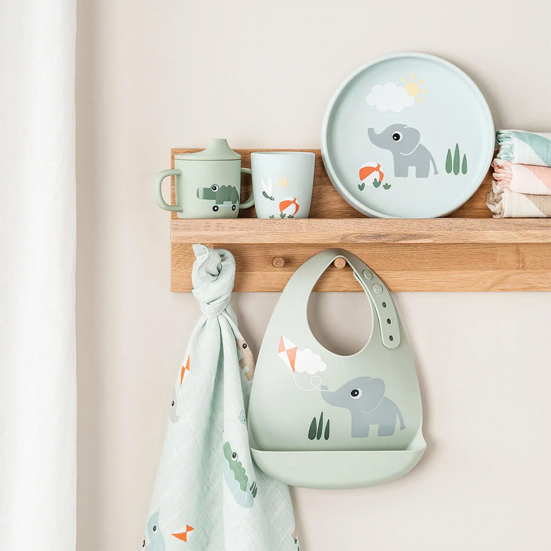 Een Done by Deer collectie van groene babyartikelen, waaronder een slabbetje, bord, bekers en een hydrofiele doek, hangt aan een houten plank.