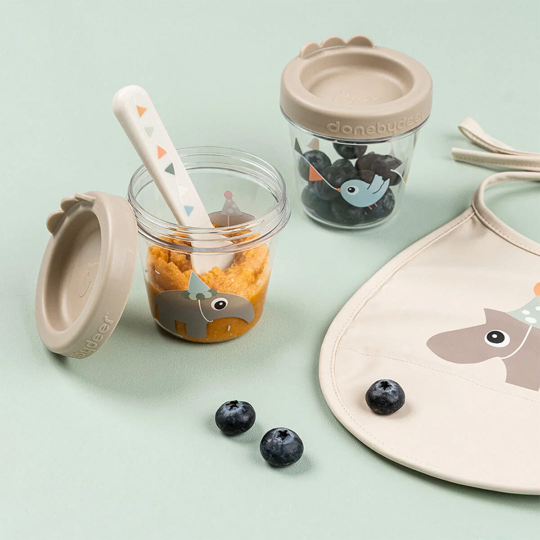 Een Done by Deer snackdoosje met babyvoeding en lepel, een gesloten snackdoosje met bosbessen en een bijpassend slabbetje op een groene ondergrond.