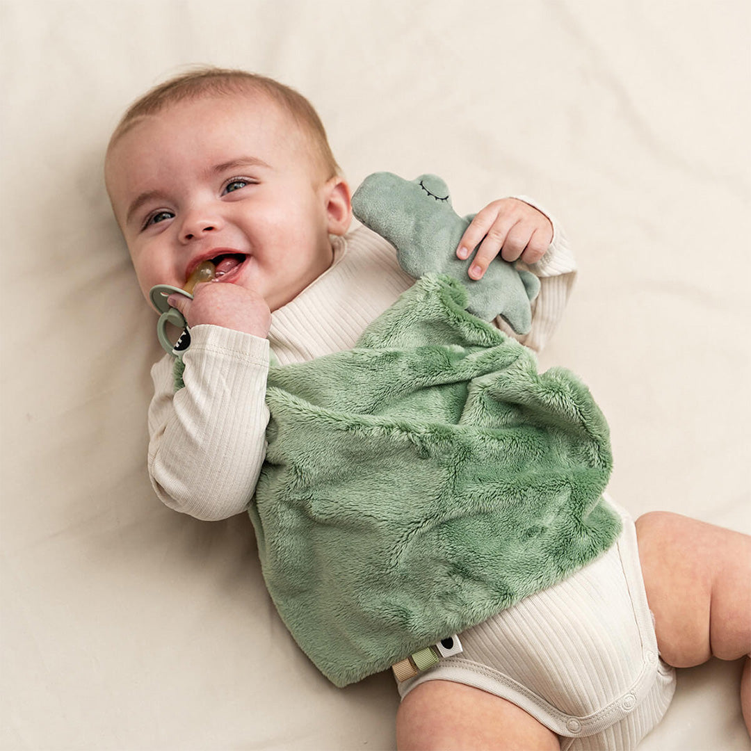 Een lachende baby met speen in de mond houdt de Done by Deer groene krokodil babyspeendoek vast.