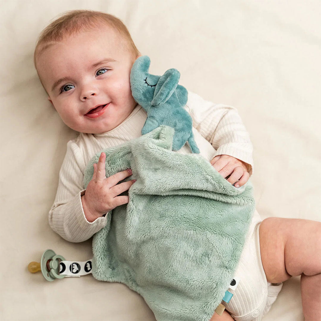 Een lachende baby ligt op de rug met de Done by Deer groene Elphee speendoek en een speen.