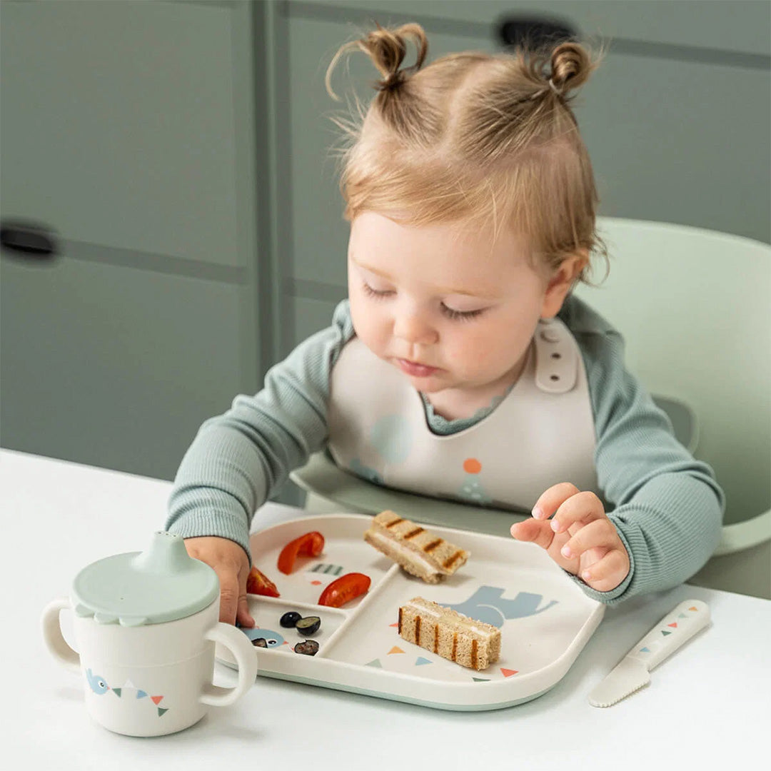 Een baby zit aan tafel met een done by deer tuitbeker en een bijpassend bord met eten.