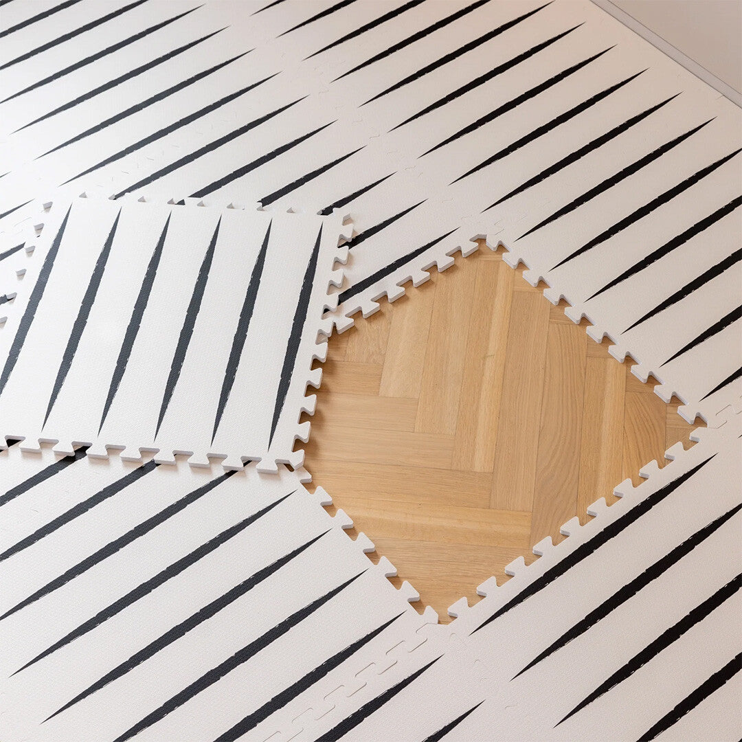 Detail van de eeveve foam speelmat met ecru en zwart geometrisch patroon, waarbij een stuk is verwijderd en een houten vloer zichtbaar is.