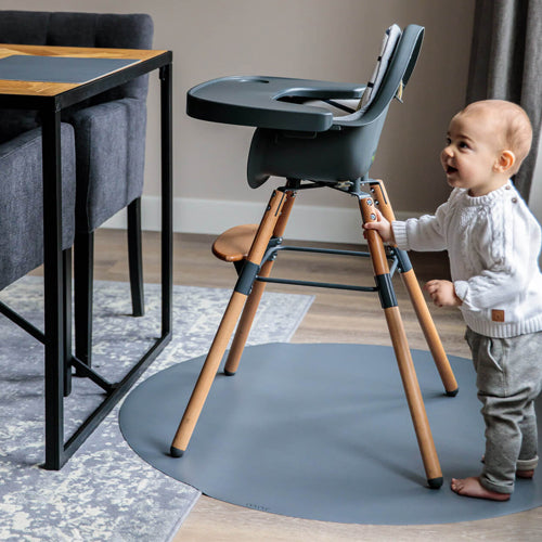 Een baby staat naast een kinderstoel op een ronde grijze eeveve knoeimat.