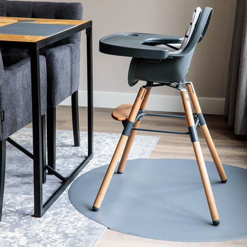 Een kinderstoel staat op een ronde grijze eeveve knoeimat in een eetkamer.