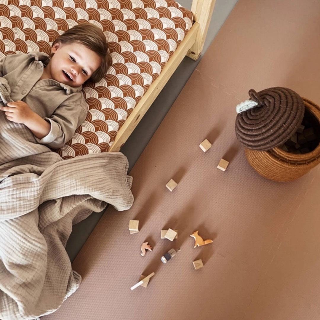 Een lachend kind ligt in bed naast de eeveve foam speelmat met houten blokken en speelgoed.