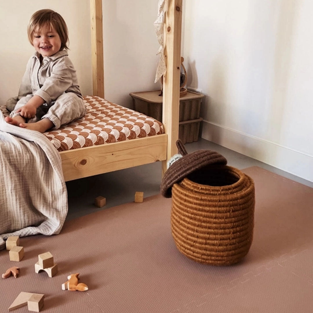 Een lachend kind zit op bed naast de eeveve foam speelmat met houten blokken en een opbergmand.