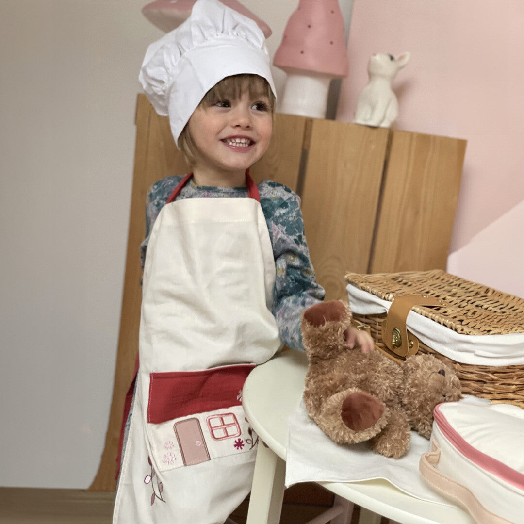 Een lachend kind draagt het Egmont Toys kinderschort met huisje en een witte koksmuts, spelend met een teddybeer.
