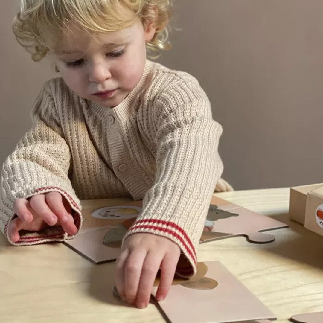 Een peuter legt een stukje van de Marcel kinderpuzzel met een beer op een houten tafel.