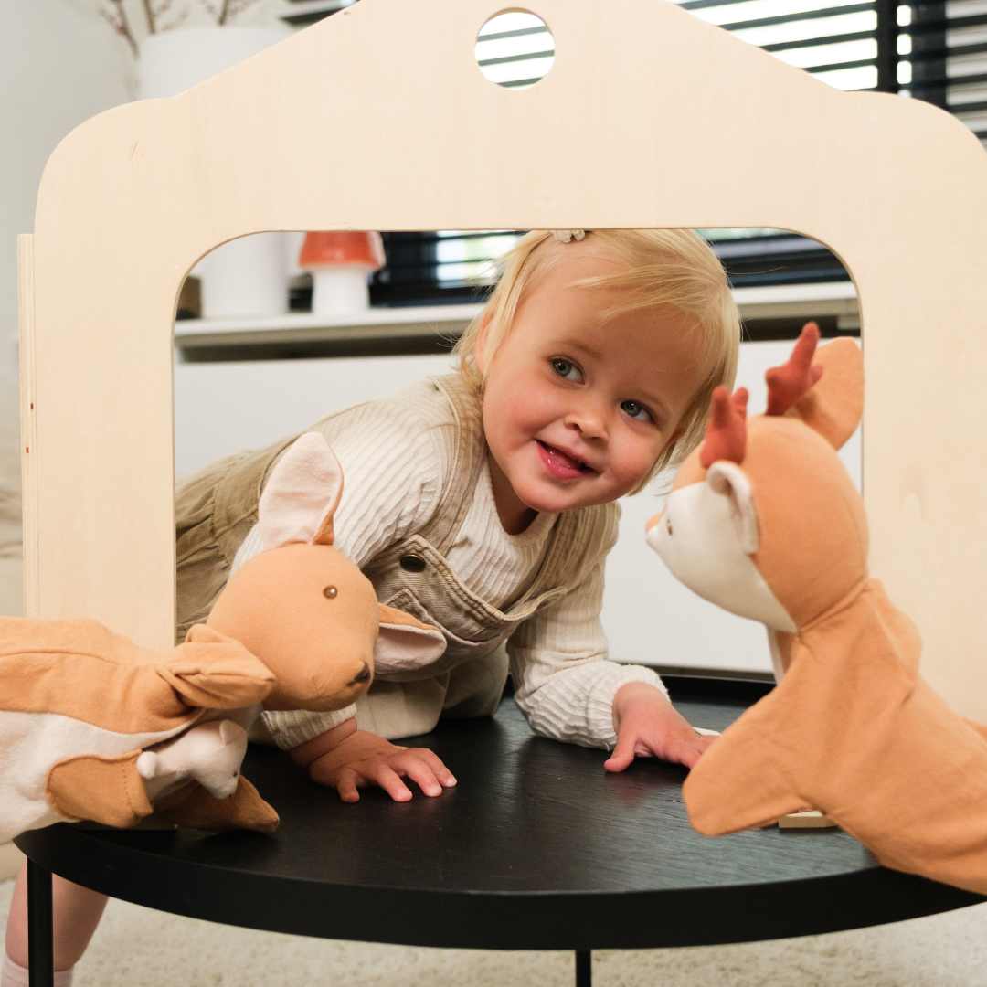 Een lachend kind speelt met twee Egmont Toys kangoeroe handpoppen achter een houten poppenkast.